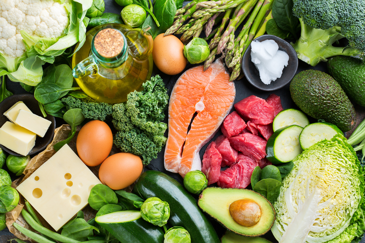 A spread of ketogenic foods with salmon in the center surrounded by red meat, green vegetables, eggs, and oil.
