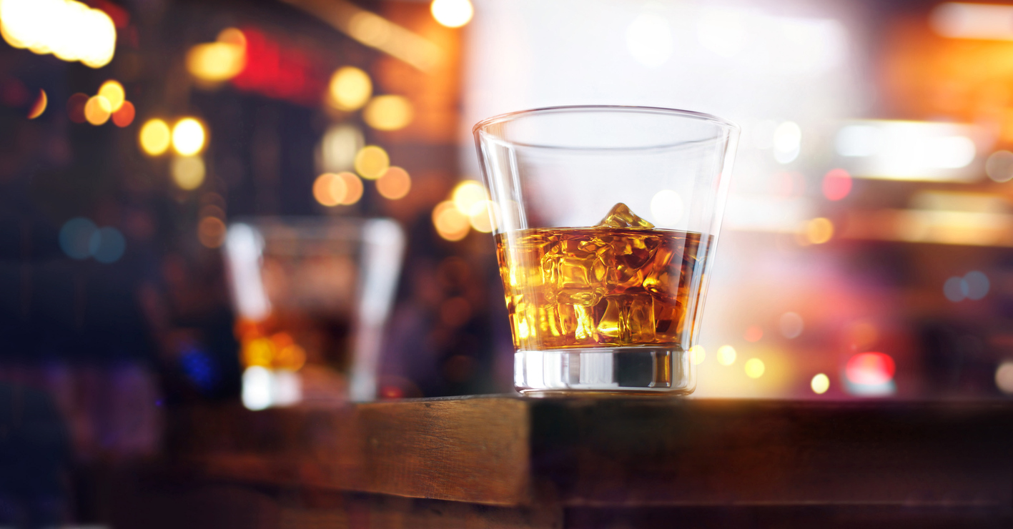 A tumbler of whiskey with ice on the corner of a wooden table with soft lighting in the background.