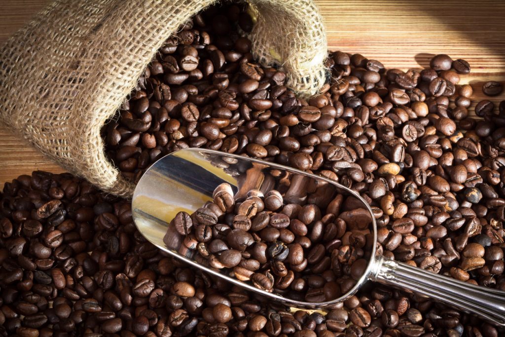 A burlap sack of coffee beans spilled onto a wooden surface with a metal scoop filled halfway with coffee beans.