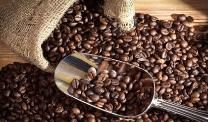 A burlap sack of coffee beans spilled onto a wooden surface with a metal scoop filled halfway with coffee beans.