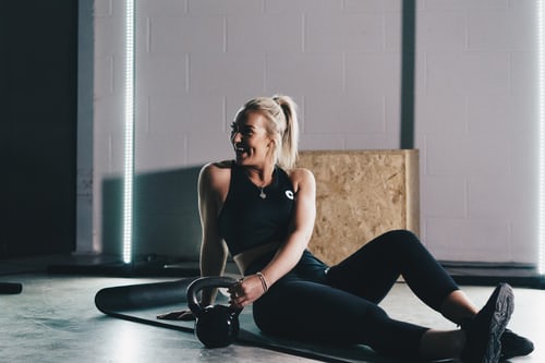 A woman sitting on a black workout mat and twisting to hold a weight next to herself.